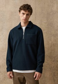 Tmavomodrý fleecový pulóver s polovičným zipsom, dlhými rukávmi a náprsným vreckom na ľavej strane. Mäkkučká textúra, voľný strih.