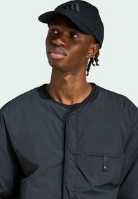 Čierna ľahká bomber bunda s hladkým povrchom, bočné vrecko so zapínaním na gombík, nosená s čiernym šiltom zladeným s logom.