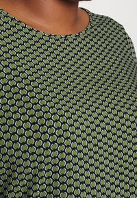 Nära bild av en person som bär en svart topp med ett grönt och vitt cirkulärt mönster och rund halsringning.