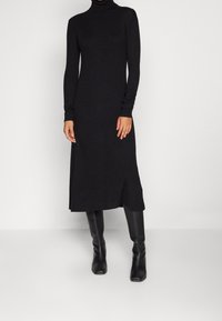 Abito nero a collo alto con costine, maniche lunghe e spacchi laterali, realizzato in tessuto morbido, silhouette aderente, fino al ginocchio, abbinato a stivali neri al ginocchio.