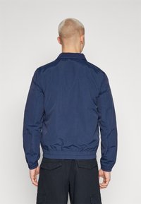 Mornar plavi bomber jakna s ovratnikom, elastičnim manžetama i pojasom. Glatka, sjajna tkanina s minimalnim detaljima šivanja.