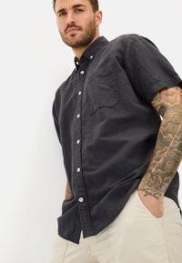 Donkergrijze kortarmige button-up shirt met een borstzak en lichte knopen, gecombineerd met beige broeken. De stof heeft een licht gekreukt textuur.