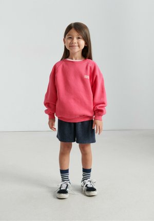 Felpa rosa con maniche raglan, taglio casual e stampa bianca "vintage". Abbinata a pantaloncini in spugna blu navy e scarpe nere con suole bianche.