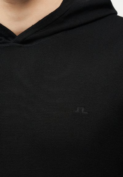 Κοντινό πλάνο ενός ατόμου που φοράει μαύρο πλεκτό πουλόβερ με κουκούλα, με ένα μικρό, διακριτικό κεντημένο λογότυπο στο στήθος.