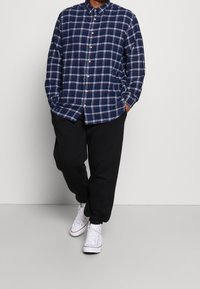 Camicia button-up a quadri blu navy con dettagli bianchi e rossi, maniche lunghe, abbinata a pantaloni tuta neri e sneakers bianche.