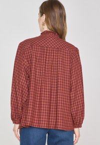 Camicia a maniche lunghe in tessuto a quadri ruggine e arancione, presenta un retro plissettato, colletto alto e polsini elasticizzati.