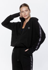 Czarna bluza z kapturem na zamek, wykonana z miękkiego materiału, charakteryzująca się luźnym krojem, przednimi kieszeniami i akcentem z logo. Detale na rękawach zawierają kontrastową grafikę.