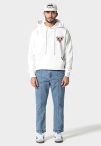 Biała bluza z kapturem z przednią kieszenią i kolorowym haftowanym wzorem. Jasnoniebieskie dżinsy z pionowymi szwami, w połączeniu z czarno-białymi trampkami.