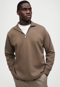 Καφέ φούτερ με φερμουάρ στον λαιμό, κατασκευασμένο από μείγμα βαμβακιού, με μακριά μανίκια και άνετη γραμμή. Φορεμένο πάνω από ένα λευκό μπλουζάκι, με απλή υφή.