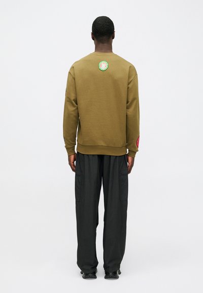 Ελιόχρωμο βαμβακερό φούτερ με στρογγυλή λαιμόκοψη, ριμπ μανσέτες και κυκλικό σήμα στην πλάτη. Συνδυάζεται με φαρδιά μαύρα cargo παντελόνια.