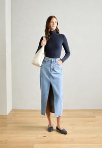 Tricou elegant cu guler înalt de culoare bleumarin, fustă maxi din denim de un albastru deschis cu o deschidere frontală și pantofi flats bleumarin; purtând o geantă de umăr de culoare crem.