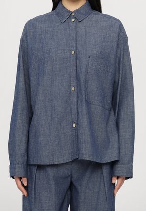 Nainen pukeutuneena löysähköön nappikiinnitteiseen farkkupaitaan, jossa on rintatasku, ja samaan sarjaan kuuluviin leveälahkeisiin farkkuihin, seisoo yksivärisen taustan edessä.