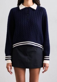 Mörkblå stickad tröja med kabelmönster, vit krage, svarta knappar och randig ribbad nederkant och ärmslut. Matchad med en svart kjol.