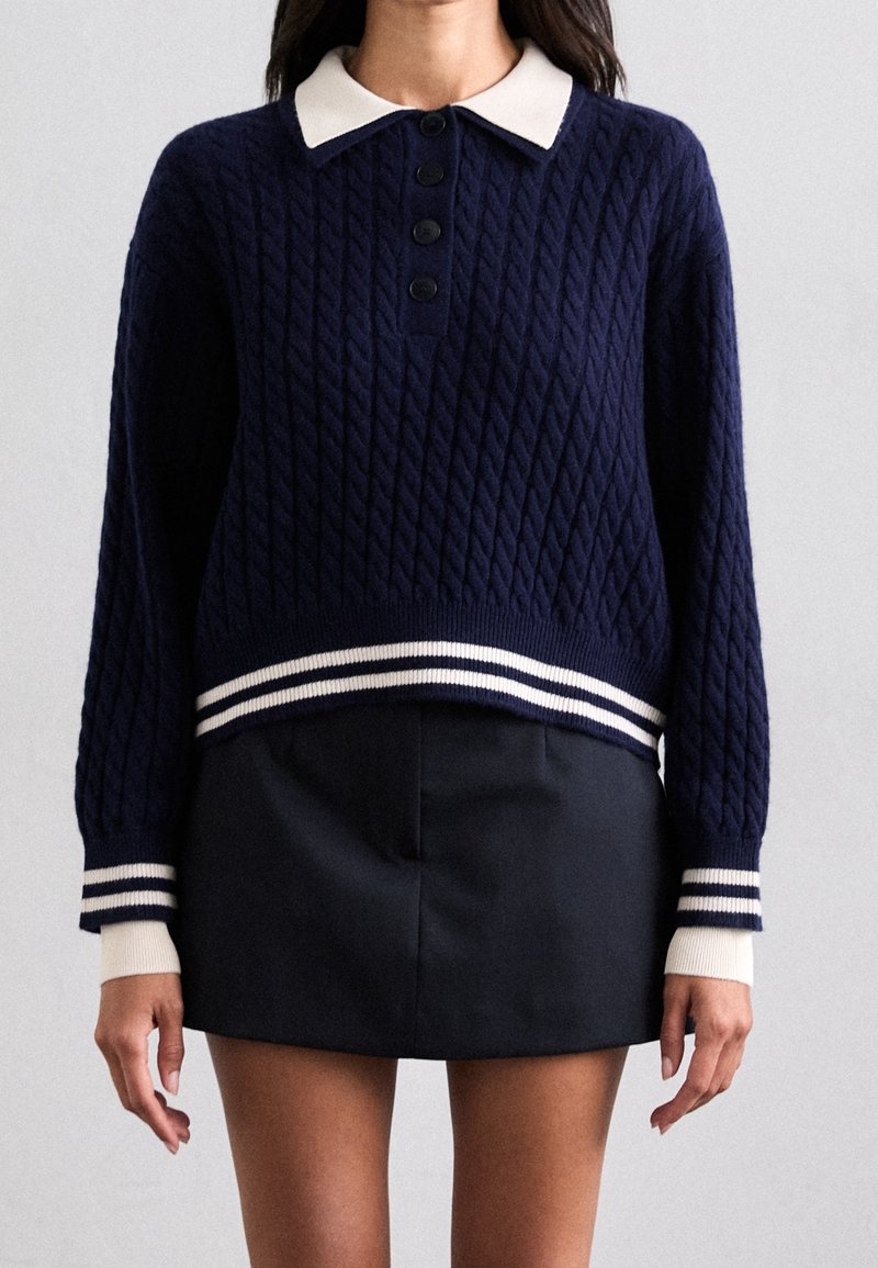 Mörkblå stickad tröja med kabelmönster, vit krage, svarta knappar och randig ribbad nederkant och ärmslut. Matchad med en svart kjol.