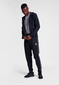 Zwarte sportjacket met capuchon, grijs T-shirt en zwarte trackpants met chevronpatroon. Zwarte sportschoenen maken de outfit compleet.