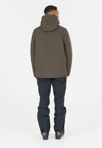 Olivgrüne wasserdichte Jacke mit Kapuze, gerade geschnitten, mit nahtlosem Design und elastischen Bündchen, kombiniert mit schwarzen Skihosen und Stiefeln.