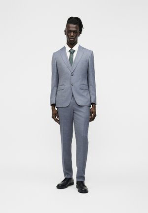 Άνδρας με ντραντλόκς φορώντας ανοιχτό γκρι κοστούμι με δύο κουμπιά, λευκό πουκάμισο, πράσινη γραβάτα και μαύρα παπούτσια, στέκεται μπροστά σε λευκό φόντο.