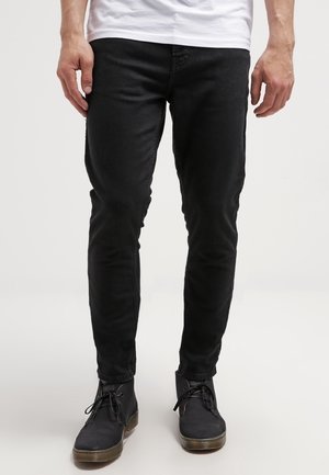 Strak zittende zwarte jeans gedragen door een persoon met zwarte veterlaarzen en een wit overhemd, staand tegen een effen lichte achtergrond.