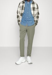 Chaqueta a cuadros verde y blanca sobre una camisa de denim azul claro, combinada con pantalones verde oliva y zapatillas blancas gruesas.