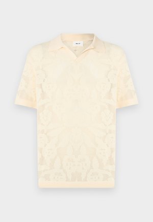 Polo de punto color crema con mangas cortas, patrón floral de malla y cuello puntiagudo, exhibido sobre un fondo liso.