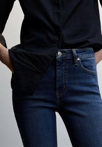 Person wearing dark blue jeans and black long-sleeve shirt, with one hand tucked into jeans pocket, shown from waist to mid-thigh.