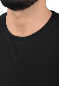 Fekete, kerek nyakú pulóver ribbed textúrával és egy apró V-varrással a nyakkivágás alatt. Az anyag lágy és kissé elasztikusnak tűnik.