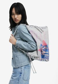 Leichter grauer Canvas-Zugbeutel mit buntem Grafikdruck, ausgestattet mit Ösen und Schnürsenkeln zum Verschließen. Das Modell trägt eine gestreifte Jacke.