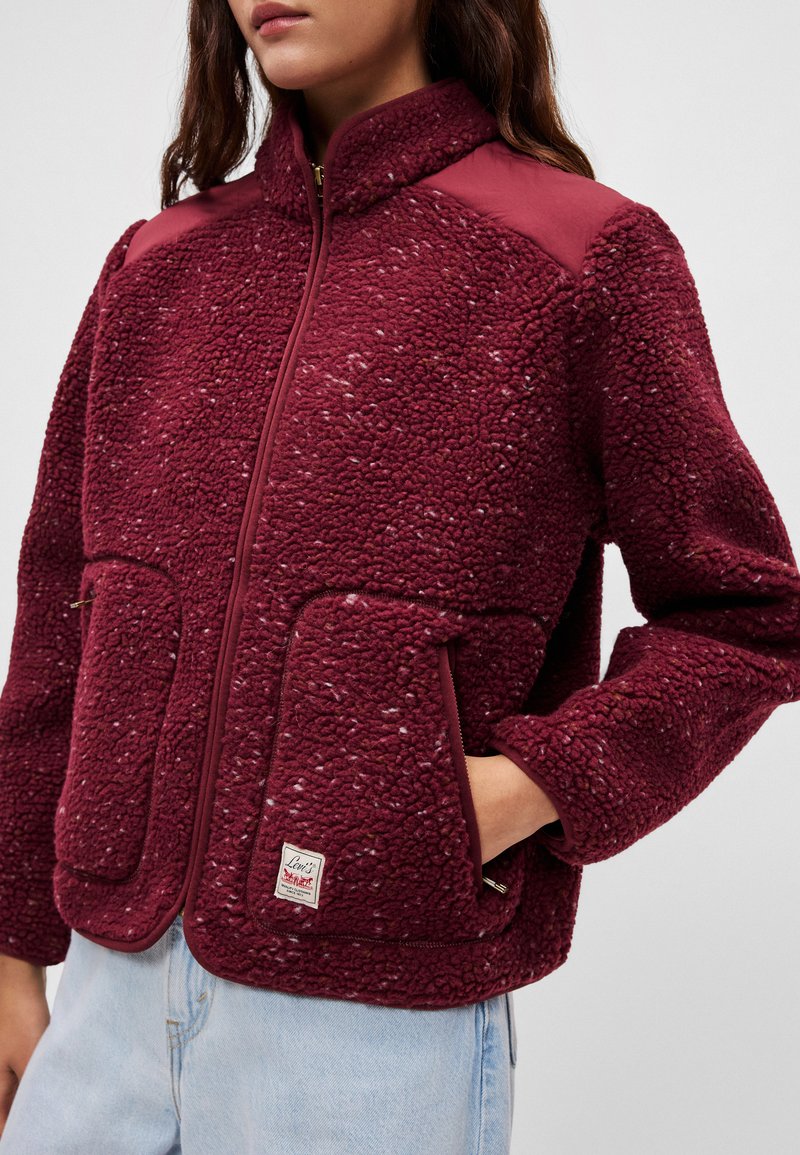 Burgundy fleece jacket with structured shoulders, zip front, two front pockets, and logo patch. Soft, textured fabric with ribbed accents.