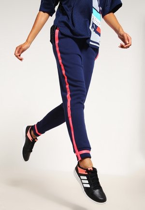 Person, der går, iført mørkeblå joggingbukser med lyserøde sidelister, sorte og lyserøde sneakers og en mørkeblå mesh-top med farverige detaljer.