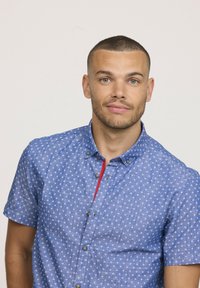 Chemise bleu à manches courtes avec des motifs floraux blancs, bouton devant et accent rouge le long de la patte de boutonnage, en tissu léger.