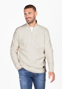 Suéter de punto color crema con media cremallera, que presenta un patrón texturizado y una pequeña etiqueta de logo marrón cerca del dobladillo.