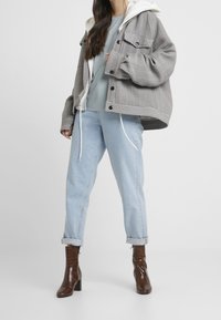 Šedá a černá kostkovaná oversized bunda, světle modrý svetr, ohrnuté světlé džínové kalhoty a hnědé boty na podpatku s texturovaným povrchem.