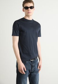 T-shirt azzurro navy a maniche corte in tessuto morbido, abbinato a jeans in denim blu scuro. Gli accessori includono occhiali da sole neri stretti e una cintura nera.