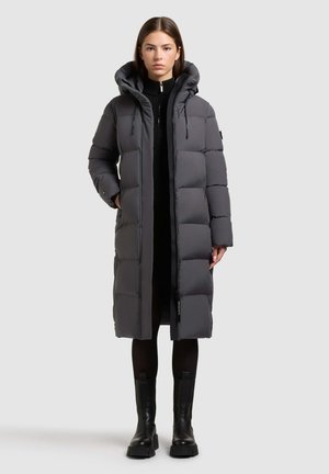 Graue Puffjacke mit gestepptem Design, übergroßer Kapuze und Reißverschluss. Getragen über einem schwarzen Kleid und kombiniert mit schwarzen klobigen Stiefeletten.