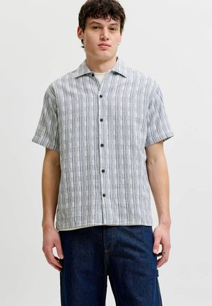 Jonge man draagt een gestreept, kort mouw, met knopen overhemd over een witte top en donkerblauwe jeans, staand tegen een effen achtergrond.