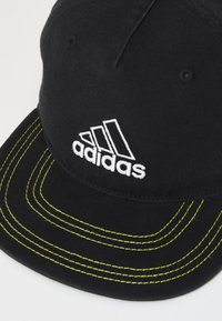 Casquette noire avec le logo blanc d'Adidas. Présente des coutures jaunes contrastées le long de la visière. Fabriquée en tissu avec une texture lisse et une forme structurée.