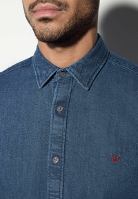Donkerblauwe denim overhemd met knopen en een casual kraag; voorzien van zwarte knopen en een klein rood geborduurd logo aan de linkerkant.