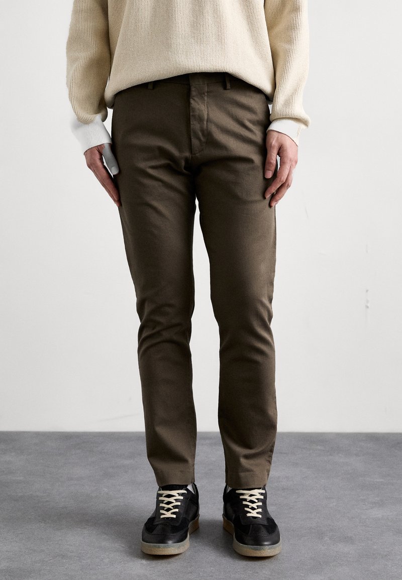 Lysbeige ribbet sweater, brune tætsiddende bukser og sorte sneakers med beige snørebånd. Skoene har en tekstureret sål og subtile sorte accenter.