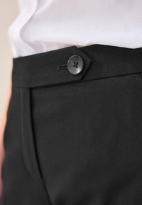 Black tailored trousers feature a smooth fabric, a flat front design, and a single black button fastening, with neat stitching details.