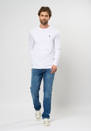 Tricou alb cu mâneci lungi și un mic logo bleumarin, asortat cu blugi albaștri și adidași albi, pe un fundal simplu.