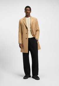 Cappotto di lana color tan con revers a punta, design doppiopetto e quattro bottoni, abbinato a un maglione crema e pantaloni neri larghi.