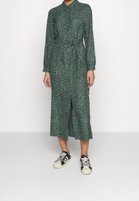 Robe midi verte avec un motif de feuilles, devant boutonné, manches longues, taille cintrée avec un lien, associée à des baskets noir et blanc.