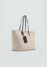 Sac fourre-tout en toile beige avec des anses en cuir noir et une étiquette rectangulaire. Présente une surface texturée et un design ouvert.