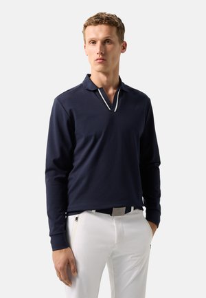 Mand iført mørkeblå langærmet polo med hvidstribet krave og hvide bukser, stående med den ene hånd i lommen mod en ensfarvet baggrund.