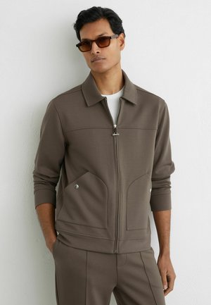 Muž v hnedej vetrovke so zipsom a veľkými prednými vreckami a v zodpovedajúcich nohaviciach stojaci pred bielou textúrovanou stenou.