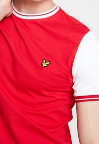 Camiseta roja de manga corta con un cuello y acentos en las mangas blancos, con un pequeño logotipo de un pájaro amarillo bordado en el pecho.