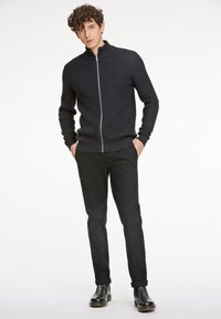 Suéter negro de punto con cremallera y un patrón texturizado, combinado con pantalones negros ajustados y botas negras de tobillo. Diseño simple y entallado.