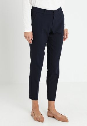 Woman wearing navy slim-fit ankle pants, white long-sleeve top, and beige pointed flats with buckle detail, standing against white background.