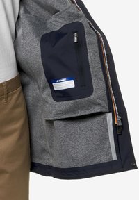 Giacca grigia con accenti blu navy, dotata di tasca con zip e foderature interne. Il materiale sembra texturizzato, con dettagli della zip a contrasto.