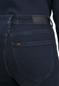 Närbild av marinblå denimjeans bakficka med Lee-märket på midjan och en liten etikett på fickkanterna.